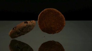Çikolata parçalı kurabiyeler, tatlı atıştırmalık ve fırın reklamı için yavaş çekimde karanlık bir cam yüzeyde düşüp zıplıyorlar.