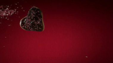 Kalp şeklinde çikolatalı çörek, pembe ve beyaz serpintiler havada uçuşuyor. Tatlı sevgililer günü hediyeleri için parlak kırmızı bir arka planda.