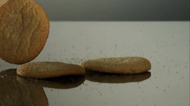 Gevrek altın şekerli kurabiyeler yavaş çekimde yansıtıcı gri bir yüzey boyunca düşüyor ve kayıyor. Küçük şeker kristalleri reklam ve gıda pazarlama içeriği için etrafa saçılmış durumda.