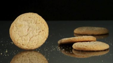 Yuvarlak, kahverengi şekerli bir kurabiye koyu bir yansıtıcı yüzeyde hızla dönüyor ve arka planda diğer kurabiyeler ve kırıntılar var.