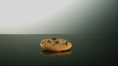 Tek bir çikolatalı kurabiyenin, yavaş çekimde yumuşak beyaz ışıltılı arka planıyla karanlık yansıtıcı bir yüzeye düşüşü.