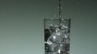 Buz küpleriyle dolu temiz bir bardağa dökülen taze maden suyu temiz, nötr gri bir stüdyonun arka planına köpükler ve su sıçratır.