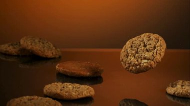 Yuvarlak pişmiş yulaflı kurabiyeler, arka planda yavaş çekimde reklamlar için sıcak turuncu bir parıltıyla koyu bir yansıtıcı yüzeyde düşüp yuvarlanıyor.