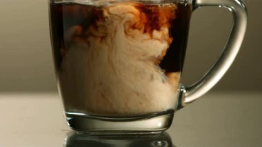 Kremalı beyaz süt şeffaf bir bardak koyu siyah kahveye dökülüyor ve sıvının içinde dönen güzel bulut desenleri oluşturuyor.