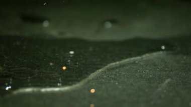 Parlayan közlerin düşük açılı makro çekimi ve ıslak, koyu dokulu bir yüzeye yavaşça yayılan altın kıvılcımlar.