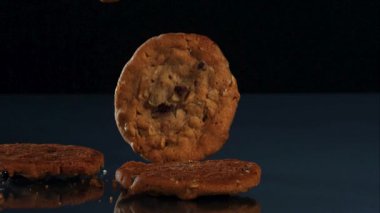 Yuvarlak yulaflı kuru üzümlü kurabiyelerden oluşan bir koleksiyon yavaş çekimde siyah bir arka plana karşı yansıtıcı mavi bir yüzeye düşüp zıplıyor.