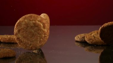 Çoklu yuvarlak tarçınlı tarçınlı kurabiye ve yulaflı kurabiyeler koyu bir yansıtıcı yüzeyde ağır çekimde koyu kırmızı arka plana doğru yuvarlanıyor.