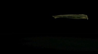 Yüz dolarlık banknot, karanlık bir arka planda yavaşça aşağı süzülüyor. Yansıtıcı bir yüzeye iniyor. Kişisel tasarrufları ve ekonomik karı temsil ediyor.