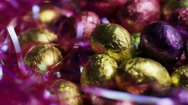 Yansımaları gösteren bir yığın içinde dönen pembe süslemelerle çevrili renkli metalik folyodan çikolatalı paskalya yumurtalarını kapatın.