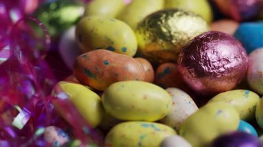 Alüminyum folyoya sarılı renkli çikolata dolu Paskalya yumurtaları ve bayram kutlamaları arka planında dönen parti flamaları.