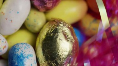Benekli pastel şeker yumurtaları ve parlak metalik folyo kaplı çikolatalar canlı pembe paskalya çimenleri arasında dönüyor neşeli bir mevsimlik arka plan oluşturuyor.