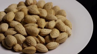 Sağlıklı atıştırma ve vejetaryen diyet kavramları için siyah arka planı olan beyaz bir tabakta yavaşça dönen büyük organik badem yığınları.