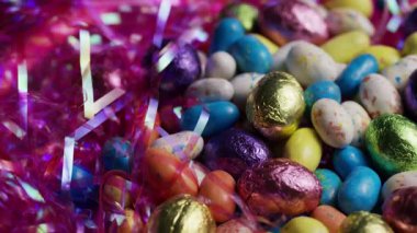 Renkli folyo çeşitleri, çikolatalı paskalya yumurtaları ve noel için parlak pembe dekoratif çimenlere yerleştirilmiş benekli jöleler.