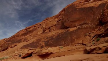 Uçsuz bucaksız bir çöl ortamında ince beyaz bulutlu, mavi gökyüzü altındaki devasa kızıl kayalık uçurumlarının alçak açılı çekimleri.