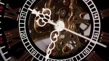Zarif bir iskelet saat yüzünün makro görüntüsü. Beyaz süslü ellerin arkasında görünen karmaşık altın dişliler büyük beyaz Roma rakamları işaretlerini geçiyor.