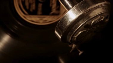 Makro perspektif, dönen siyah bir plak plağını takip ederken, eski bir gramofoncunun karmaşık metal ses kutusu ve iğne montajına odaklanıyor