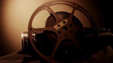 Eski moda sinema kamerası ve sıcak odaklı spot ışıklarının altında karanlık bir kumaş yüzeyinde duran ayrı bir film makarası.