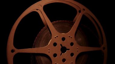 Antik metal film makarası, selüloidin dokusunu vurgulayan ılık bir yan ışıklandırmayla karanlık bir arkaplan önünde dönüyor.