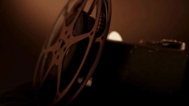 Klasik bir film kamerası ve metalik bir film makarası boyunca yavaş çekimde yumuşak altın renkli stüdyo ışıklarının altında karanlık bir yüzey üzerinde dinleniyor.