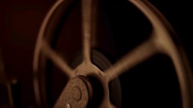 Eski bir film makarasının yakın plan çekimi. Karanlık arka planda sıcak ışıklandırması olan bir film projektörünün üzerinde hızla dönüyor.