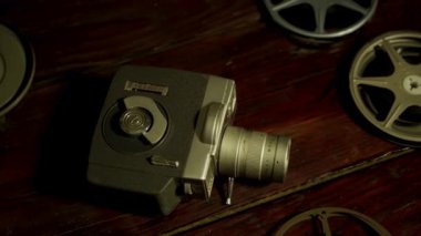 Klasik 8mm film kamerası ve birden fazla film makarası karanlık bir odada, sıcak bir ışığın altında, ahşap bir masaya saçılmış.