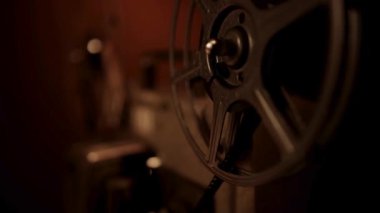 Mekanik hareketi ve karanlık odada metalik dokuları vurgulayan dönen bir film makarası olan klasik bir film projektörünün yan profili