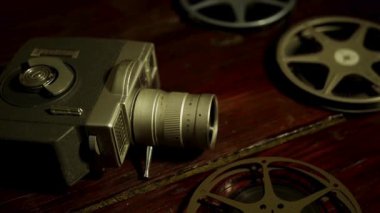 Klasik film kamerası ve film makaraları karanlık odada eski ahşap masa yüzeyinde sıcak sinematik ışıklandırma düzeneğiyle yatıyor.