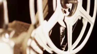 Karanlık bir stüdyoda, sıcak bir ışıkla hızla dönen eski bir projektörün üzerinde dönen bir metal film makarasının yakın çekim görüntüsü.