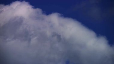 Büyük, beyaz, tüylü bulutlar, açık mavi bir gökyüzünde, güneşli bir öğleden sonra, yumuşak, doğal bir ışık altında hareket ediyorlar.