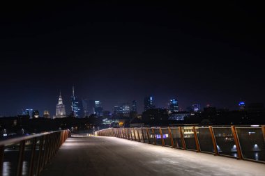 Warsaw gökdelenleri ve uzaklarda parlayan Kültür Sarayı ile modern bir yaya köprüsünden çekilen gece manzarası.