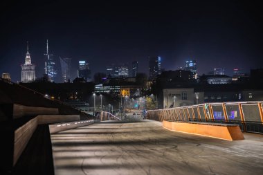 Warsaw gökdelenleri ve uzaklarda parlayan Kültür Sarayı ile modern bir yaya köprüsünden çekilen gece manzarası.