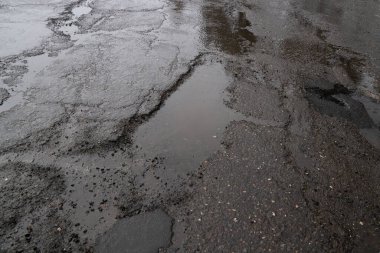 Close-up view of a severely damaged asphalt road covered in cracks, deep potholes filled with rainwater, and scattered gravel, showcasing poor road maintenance on a gloomy, overcast day.