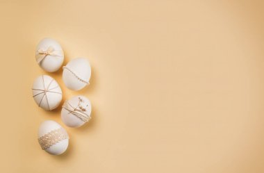 Kurdele ve dantel kullanılarak farklı şekillerle süslenmiş beş yumurta. Renkler yumuşak ve aydınlık, Paskalya için şenlikli bir atmosfer yaratıyor..