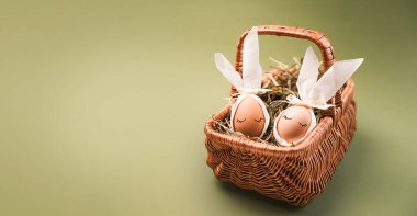 Tavşan gibi görünmek için tasarlanmış iki dekoratif Paskalya yumurtası samanla dolu bir sepetin içinde oturuyor. Ayar, Paskalya ve kutlamanın neşeli temasını vurguluyor.