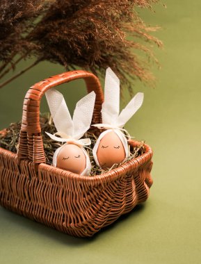 Tavşan kulaklı iki Paskalya yumurtası samanla dolu bir sepette oturur. Ayarlar yeşil bir arkaplan içeriyor. Bu sahne, Paskalya geleneklerini kutlarken neşeli bir anı yakalar..