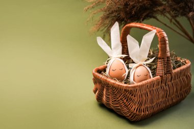 İki yumurta süslenir ve samanla dolu bir sepetin içine yerleştirilir. Beyaz fiyonkları var ve açık yeşil bir arkaplanda dizilmişler..