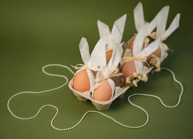 Paskalya yumurtaları bir karton içinde düzenlenir, her biri kağıtla sarılır ve kurdeleyle bağlanır. Çiçek elementleri ve danteller şenlik ortamına detay ekler. Arka plan yeşil..