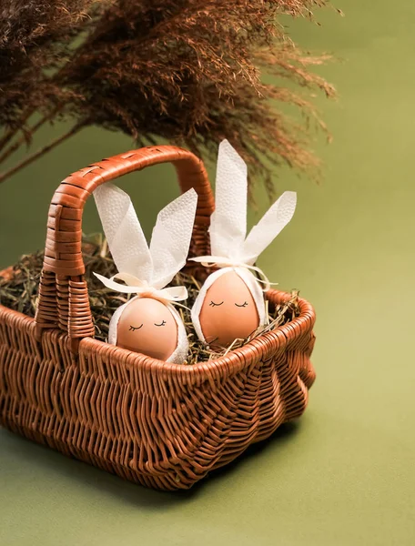 Tavşan kulaklı iki Paskalya yumurtası samanla dolu bir sepette oturur. Ayarlar yeşil bir arkaplan içeriyor. Bu sahne, Paskalya geleneklerini kutlarken neşeli bir anı yakalar..