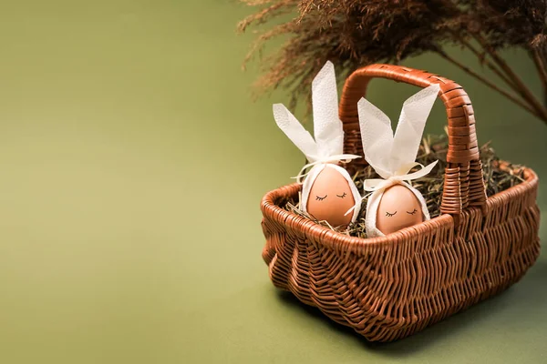İki yumurta süslenir ve samanla dolu bir sepetin içine yerleştirilir. Beyaz fiyonkları var ve açık yeşil bir arkaplanda dizilmişler..