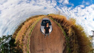 Kırsal bir mini gezegende bir arabanın yanında siyah bir adam. Siyah adam, ağaçlar, beyaz bulutlar ve yumuşak mavi gökyüzü ile 360 kamera görüntüsü.