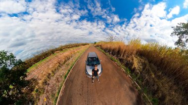 Kırsal bir mini gezegende bir arabanın yanında siyah bir adam. Siyah adam, ağaçlar, beyaz bulutlar ve yumuşak mavi gökyüzü ile 360 kamera görüntüsü.