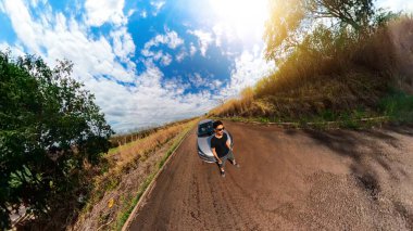 Kırsal bir mini gezegende bir arabanın yanında siyah bir adam. Siyah adam, ağaçlar, beyaz bulutlar ve yumuşak mavi gökyüzü ile 360 kamera görüntüsü.