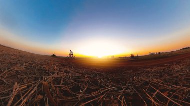 Güneş batarken kasklı ve gözlüklü siyah bir adam bisiklet sürüyor.