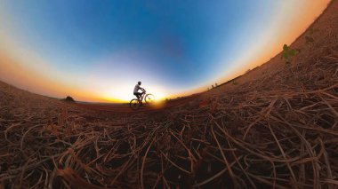 Güneş batarken kasklı ve gözlüklü siyah bir adam bisiklet sürüyor.