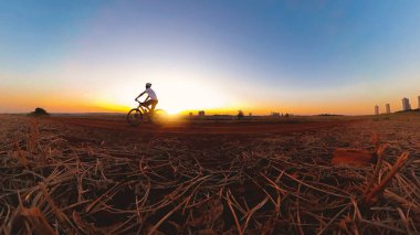 Güneş batarken kasklı ve gözlüklü siyah bir adam bisiklet sürüyor.