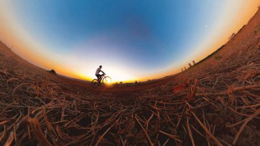 Güneş batarken kasklı ve gözlüklü siyah bir adam bisiklet sürüyor.