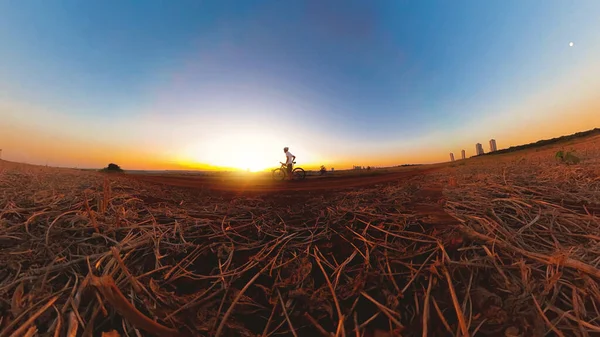 Güneş batarken kasklı ve gözlüklü siyah bir adam bisiklet sürüyor.
