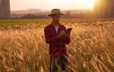 Şapkalı ve gezici tabletli çiftçi gün batımında çiftliği inceliyor. Binalar ve şehir arkaplanı bulanık.