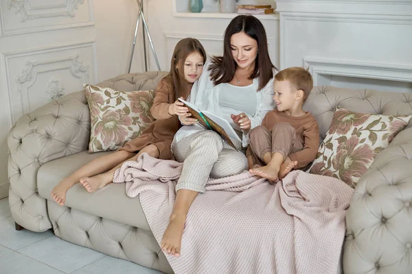 Mom reads a book to the children. A woman tells a story to a boy and a girl before going to bed. Mom daughter and son relax at home on a day off
