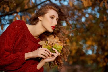 Kırmızı kazaklı kızıl saçlı genç kadın parkta yürüyor. Günbatımında gösterişli kızıl saçlı bir kadının sonbahar güzel portresi.